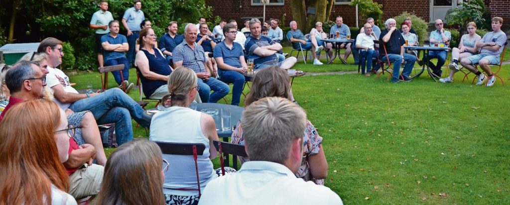 Ungefähr 40 Personen sizen auf Gartenstühlen auf einer Rasenfläche und hören interessiert zu.